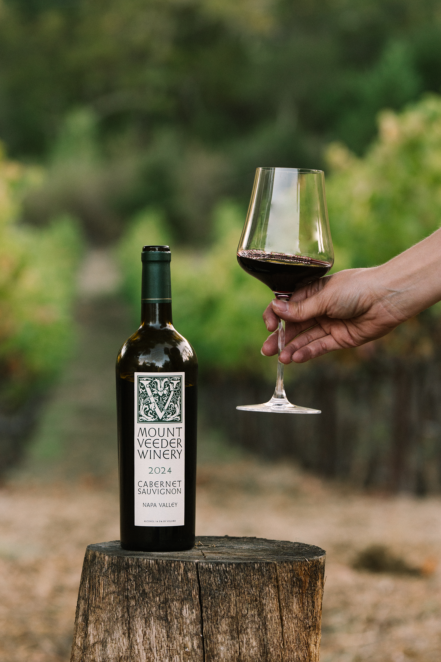 A bottle of Mount Veeder Winery 2024 Camerone Sauvignon from Sonoma Valley stands on a rustic wooden stump, with a hand holding a glass of the deep red wine against a blurred vineyard background.